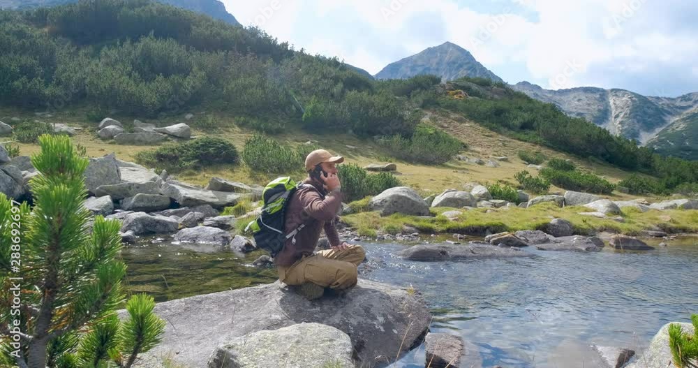 Yong male traveler in the mountains sitting near river and use cellphone Yong male traveler in the mountains sitting near river and use cellphone