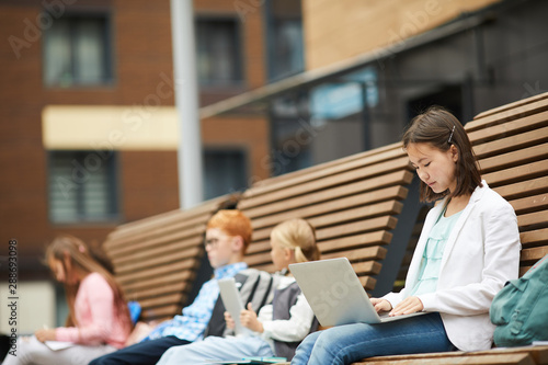 Asian schoolgirl concentrating on her study she sitting on the bench and typing on laptop computer with other school children in the background outdoors
