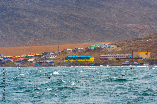 View from the sea to the Chukchi village of Enmelen. A small arctic settlement on the coast of the Bering Sea. Colorful houses on the hillside. At sea, the waves. Enmelen, Chukotka, Russian Far East. photo