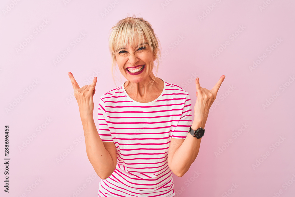 Middle age woman wearing striped t-shirt standing over isolated pink background shouting with crazy expression doing rock symbol with hands up. Music star. Heavy concept. Middle age woman wearing striped t-shirt standing over isolated pink background shouting with crazy expression doing rock symbol with hands up. Music star. Heavy concept.