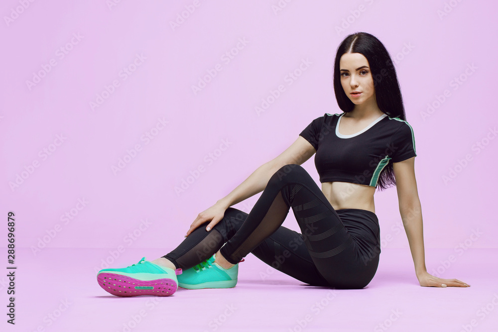 Woman in trendy sportswear relaxing after training. Young beautiful slim brunette girl in fashion black leggings, top and green sneakers posing on pink background.