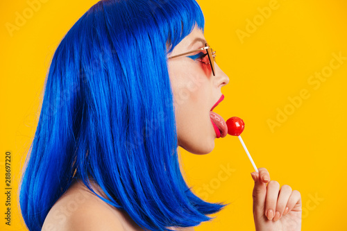Portrait in profile of glamorous nice young woman wearing blue wig and sunglasses licking lollipop