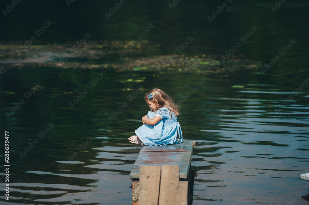 Thoughtful child is sitting on a small wooden pier on the background of the river Thoughtful child is sitting on a small wooden pier on the background of the river
