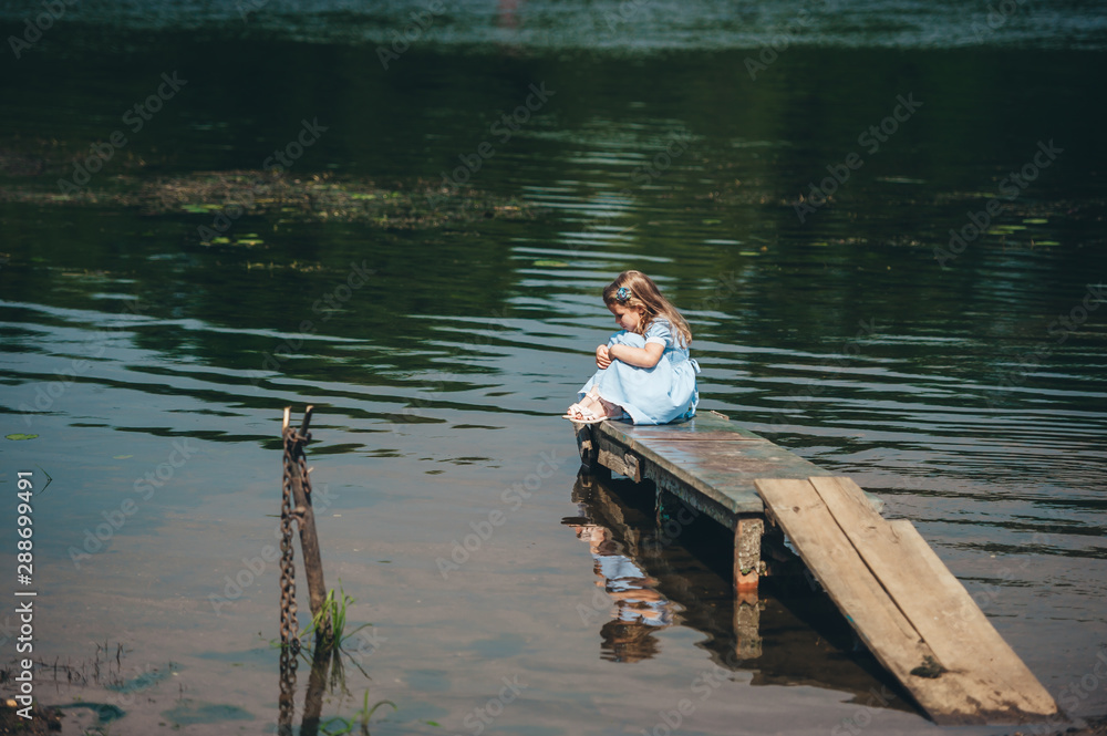 Thoughtful little girl in blue dress is sitting on a small wooden pier on the background of the lake Thoughtful little girl in blue dress is sitting on a small wooden pier on the background of the lake