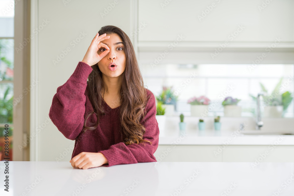 Young beautiful woman at home doing ok gesture shocked with surprised face, eye looking through fingers. Unbelieving expression. Young beautiful woman at home doing ok gesture shocked with surprised face, eye looking through fingers. Unbelieving expression.