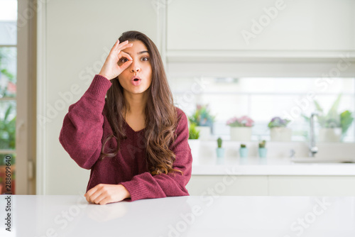 Young beautiful woman at home doing ok gesture shocked with surprised face  eye looking through fingers. Unbelieving expression.