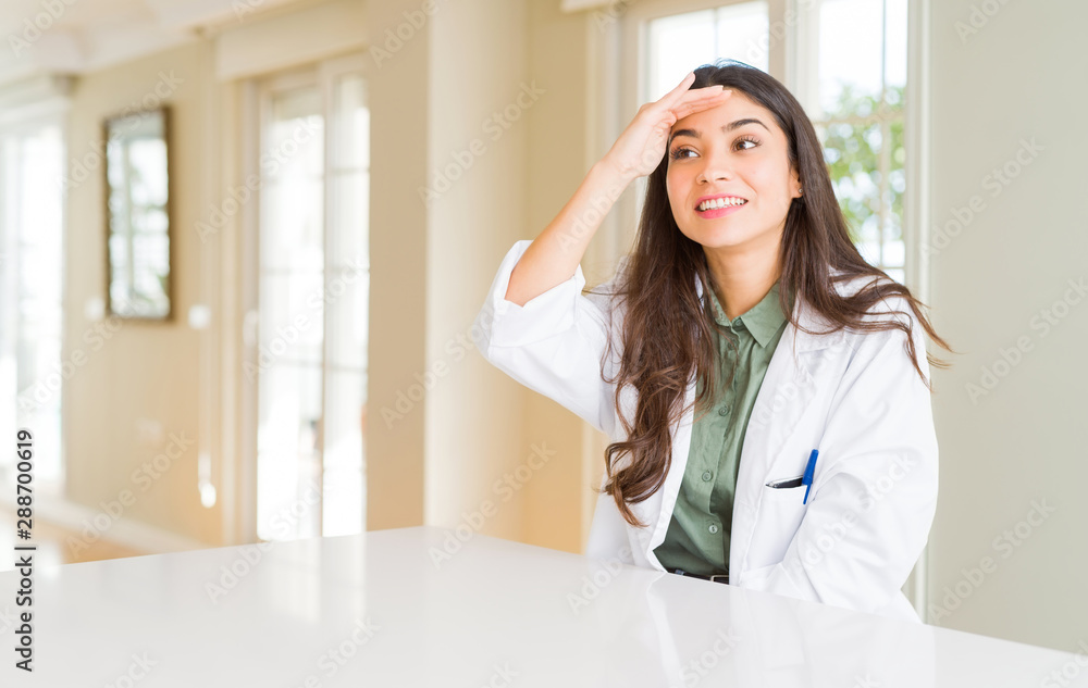 Young woman wearing medical coat at the clinic as therapist or doctor very happy and smiling looking far away with hand over head. Searching concept. Young woman wearing medical coat at the clinic as therapist or doctor very happy and smiling looking far away with hand over head. Searching concept.