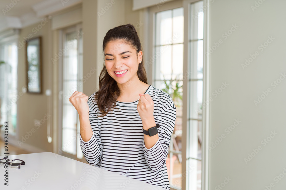 Young beautiful woman at home very happy and excited doing winner gesture with arms raised, smiling and screaming for success. Celebration concept.