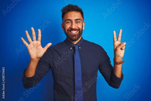 Young indian businessman wearing elegant shirt and tie standing over isolated blue background showing and pointing up with fingers number seven while smiling confident and happy.