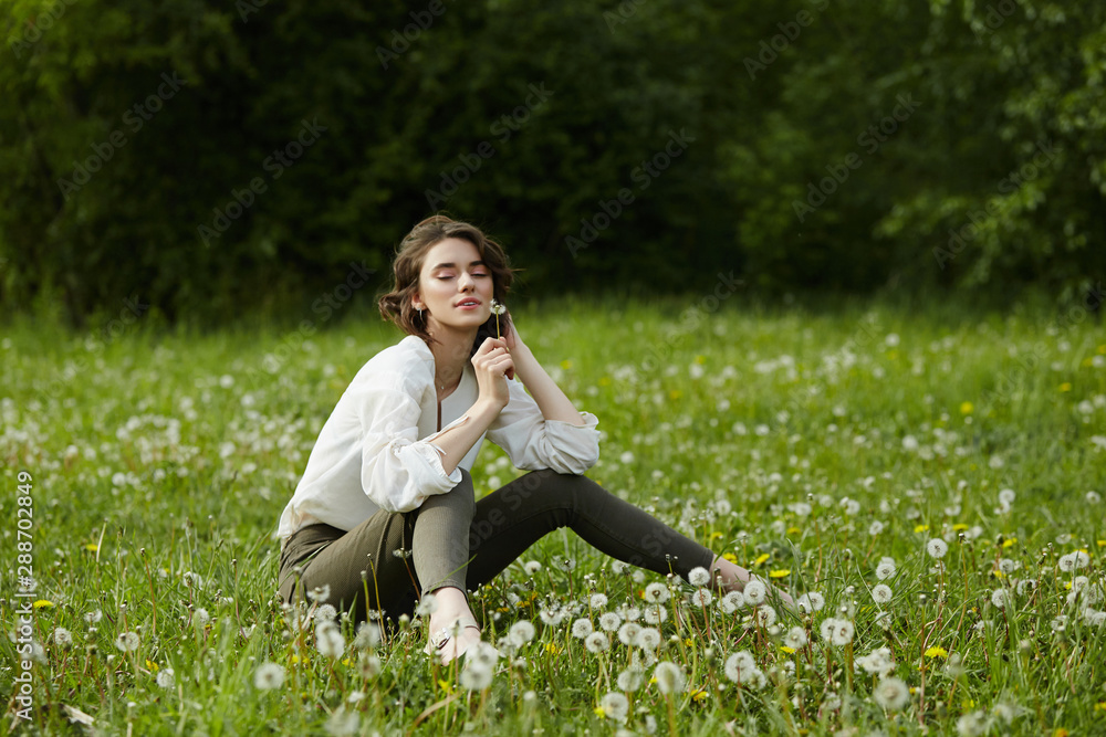Portrait of a girl sitting in a field on the spring grass among dandelion flowers. Cheerful girl enjoys Sunny spring weather. Natural beauty of a woman, natural cosmetics Portrait of a girl sitting in a field on the spring grass among dandelion flowers. Cheerful girl enjoys Sunny spring weather. Natural beauty of a woman, natural cosmetics