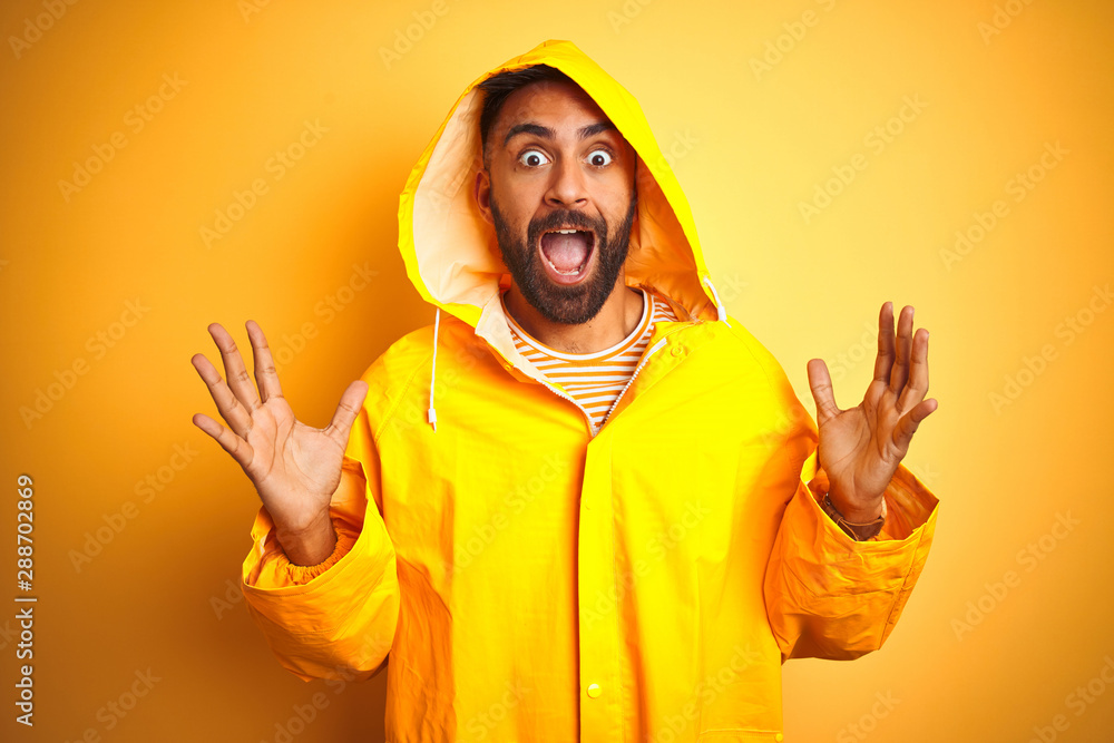 Young indian man wearing raincoat with hood standing over isolated yellow background celebrating crazy and amazed for success with arms raised and open eyes screaming excited. Winner concept Young indian man wearing raincoat with hood standing over isolated yellow background celebrating crazy and amazed for success with arms raised and open eyes screaming excited. Winner concept