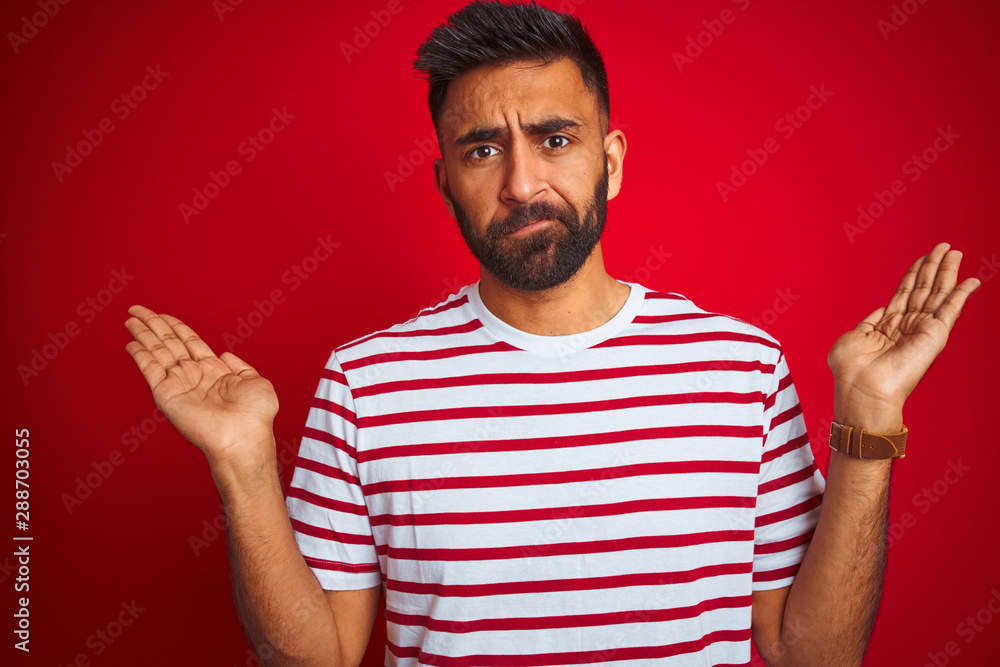 Fototapeta premium Young indian man wearing striped t-shirt standing over isolated red background clueless and confused expression with arms and hands raised. Doubt concept.