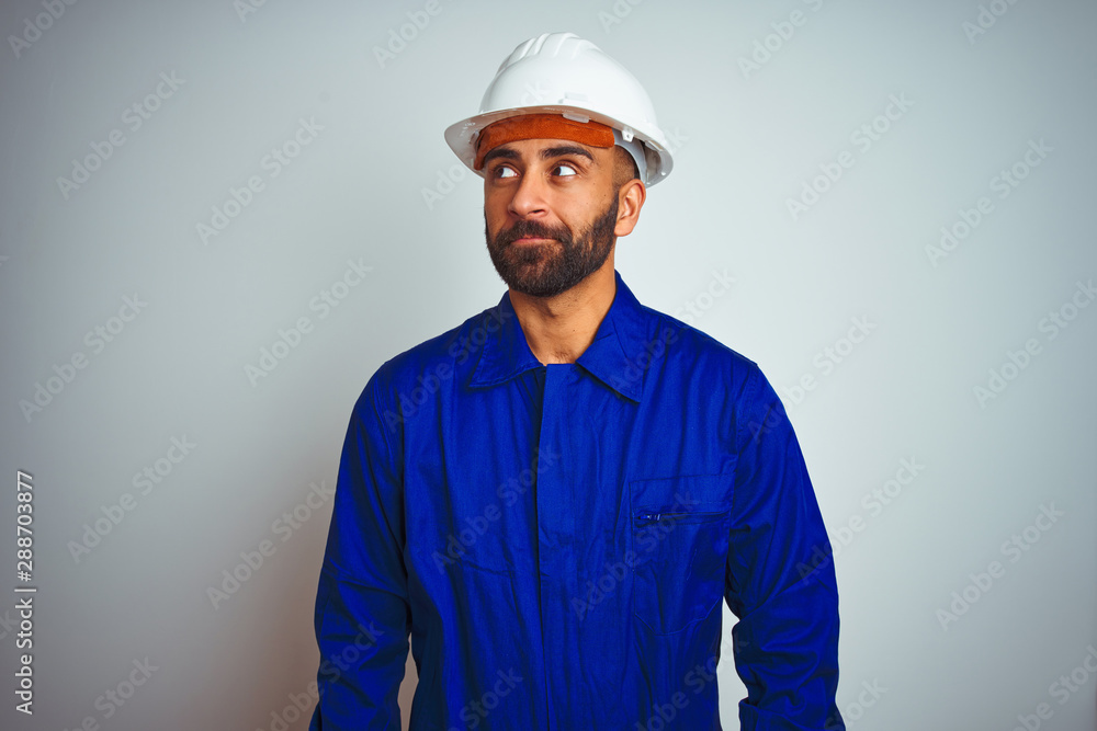Handsome indian worker man wearing uniform and helmet over isolated white background smiling looking to the side and staring away thinking.