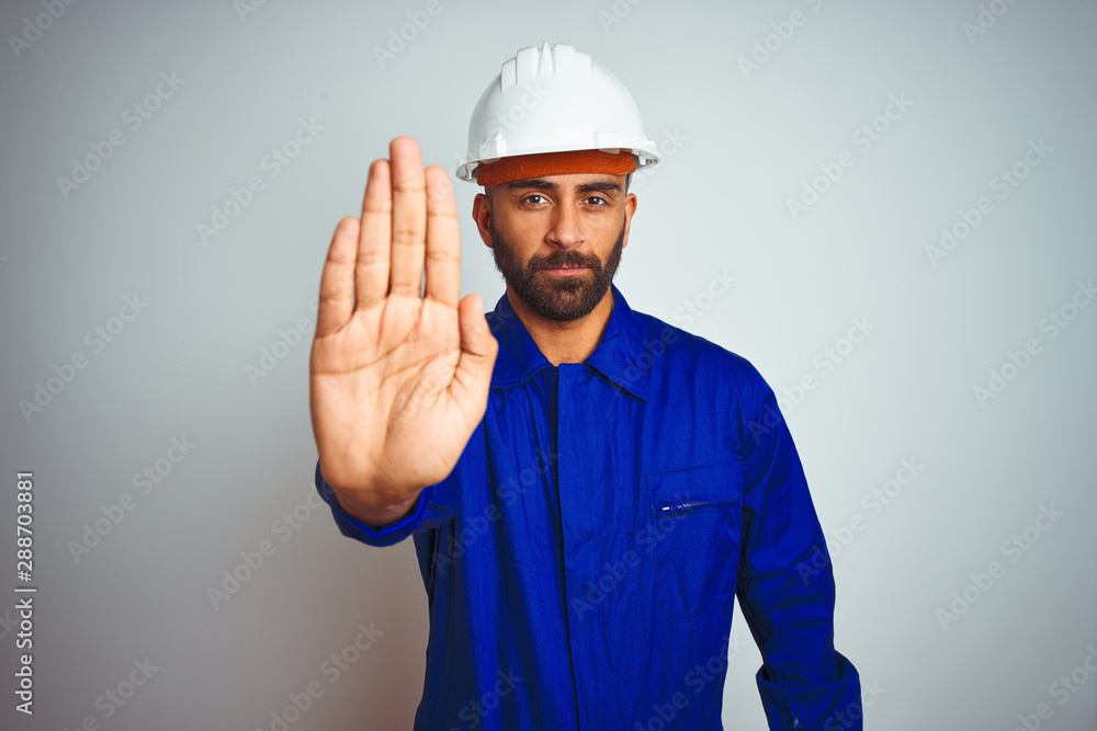 Handsome indian worker man wearing uniform and helmet over isolated white background doing stop sing with palm of the hand. Warning expression with negative and serious gesture on the face.
