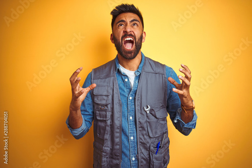 Handsome indian technician man wearing uniform over isolated yellow background crazy and mad shouting and yelling with aggressive expression and arms raised. Frustration concept.