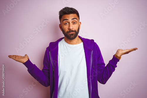 Young indian man wearing purple sweatshirt standing over isolated pink background smiling showing both hands open palms, presenting and advertising comparison and balance photo