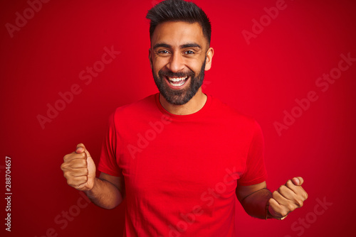 Young handsome indian man wearing t-shirt over isolated red background celebrating surprised and amazed for success with arms raised and open eyes. Winner concept. © Krakenimages.com