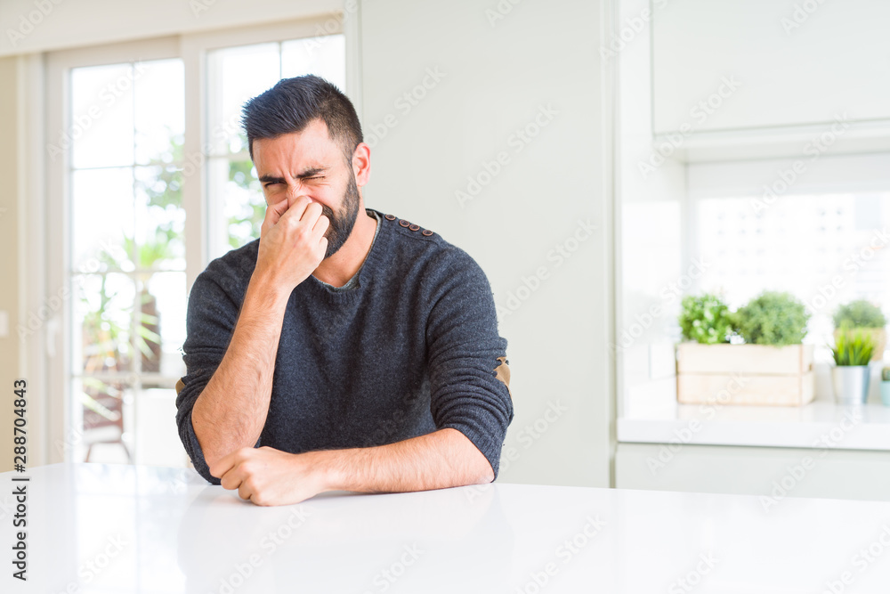 Handsome hispanic man wearing casual sweater at home smelling something stinky and disgusting, intolerable smell, holding breath with fingers on nose. Bad smells concept.