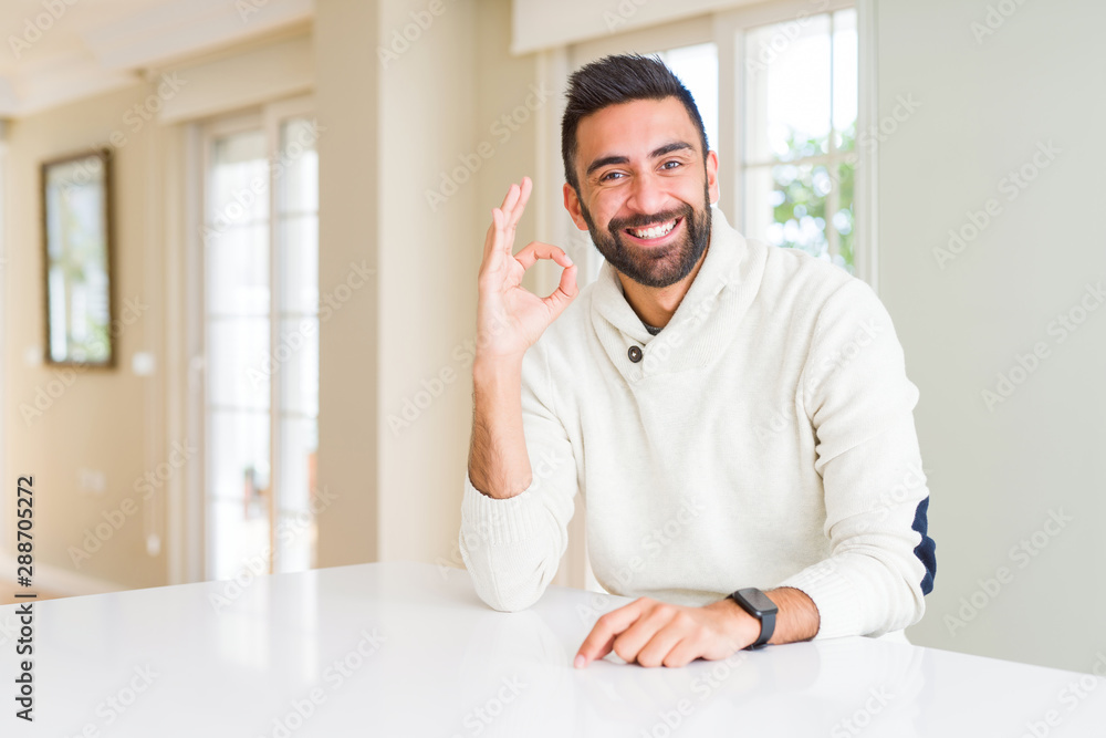 Handsome hispanic man wearing casual white sweater at home smiling positive doing ok sign with hand and fingers. Successful expression.