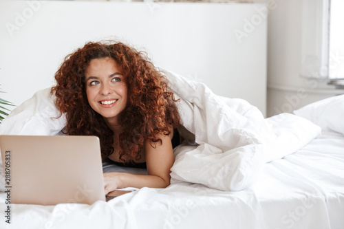 Pleased optimistic young lady in lingerie indoors in home hotel lies in comfortable bed using laptop computer.