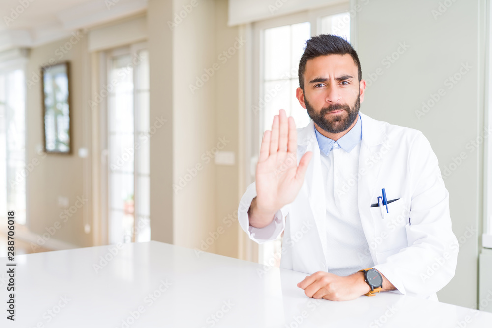Handsome hispanic doctor or therapist man wearing medical coat at the clinic doing stop sing with palm of the hand. Warning expression with negative and serious gesture on the face.