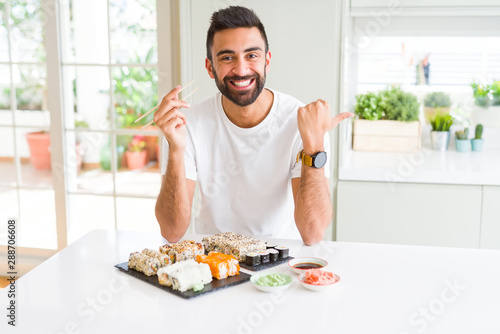 Handsome hispanic man eating asian sushi using chopsticks pointing and showing with thumb up to the side with happy face smiling