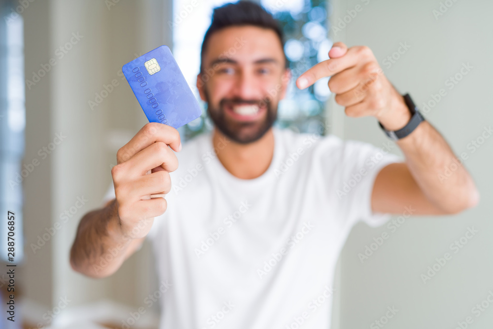 Handsome hispanic man holding credit card very happy pointing with hand and finger Handsome hispanic man holding credit card very happy pointing with hand and finger