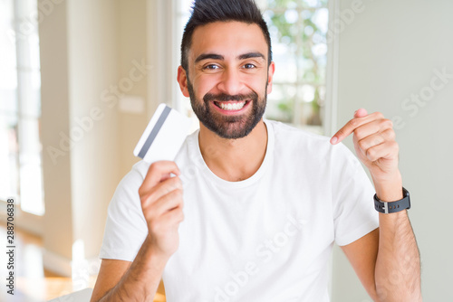 Handsome hispanic man holding credit card with surprise face pointing finger to himself