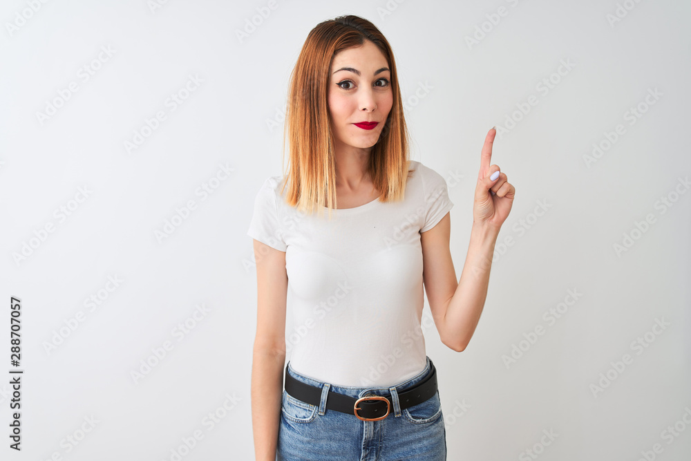 Beautiful redhead woman wearing casual t-shirt standing over isolated white background showing and pointing up with finger number one while smiling confident and happy. Beautiful redhead woman wearing casual t-shirt standing over isolated white background showing and pointing up with finger number one while smiling confident and happy.