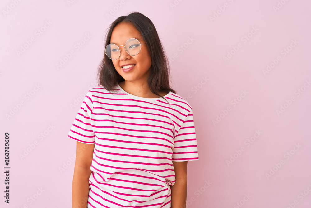 Young chinese woman wearing striped t-shirt and glasses over isolated pink background looking away to side with smile on face, natural expression. Laughing confident. Young chinese woman wearing striped t-shirt and glasses over isolated pink background looking away to side with smile on face, natural expression. Laughing confident.