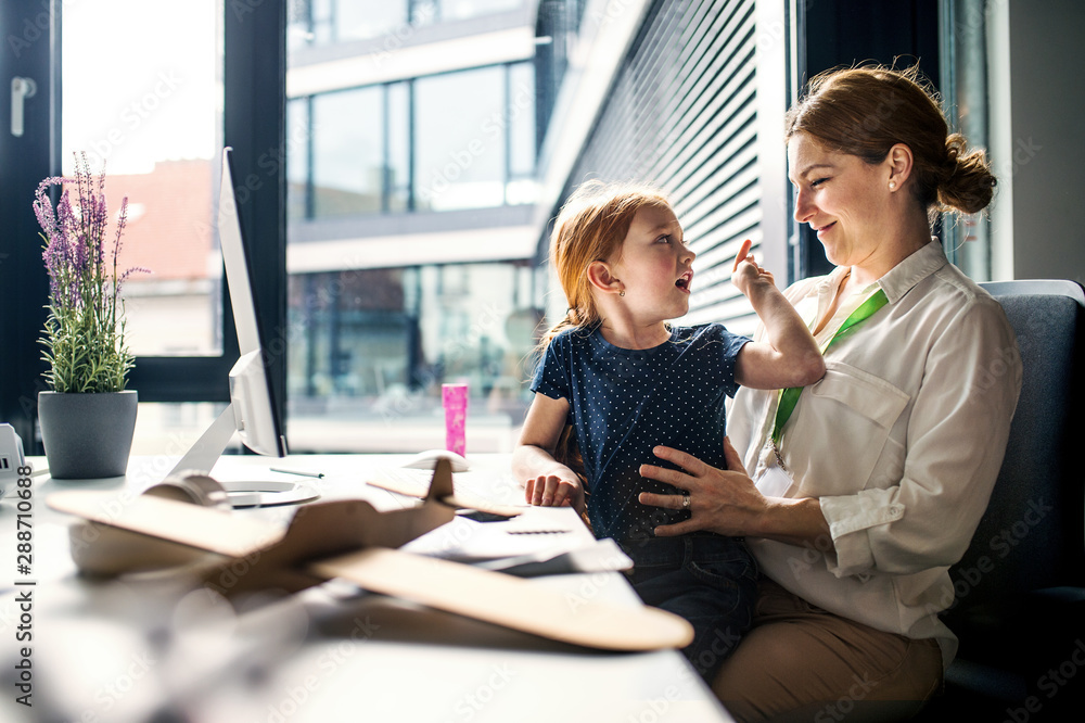 A businesswoman with small daughter sitting in an office, working. A businesswoman with small daughter sitting in an office, working.