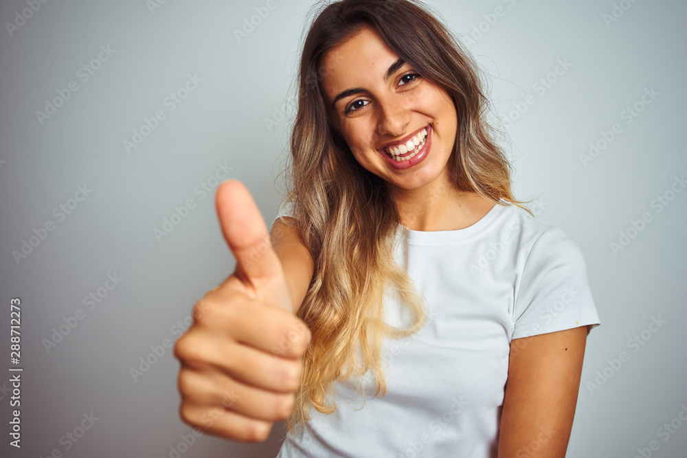 Young beautiful woman wearing t-shirt over white isolated background happy with big smile doing ok sign, thumb up with fingers, excellent sign Young beautiful woman wearing t-shirt over white isolated background happy with big smile doing ok sign, thumb up with fingers, excellent sign