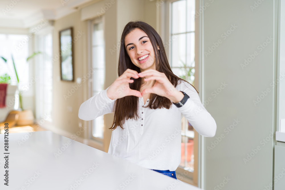 Beautiful young woman sitting on white table at home smiling in love showing heart symbol and shape with hands. Romantic concept. Beautiful young woman sitting on white table at home smiling in love showing heart symbol and shape with hands. Romantic concept.