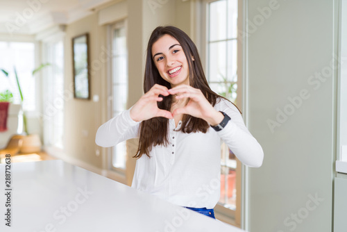 Beautiful young woman sitting on white table at home smiling in love showing heart symbol and shape with hands. Romantic concept.