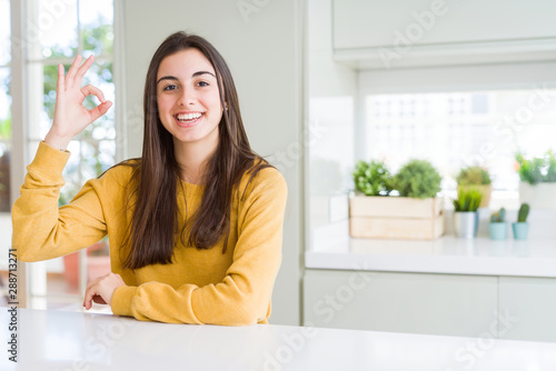 Beautiful young woman wearing yellow sweater smiling positive doing ok sign with hand and fingers. Successful expression.