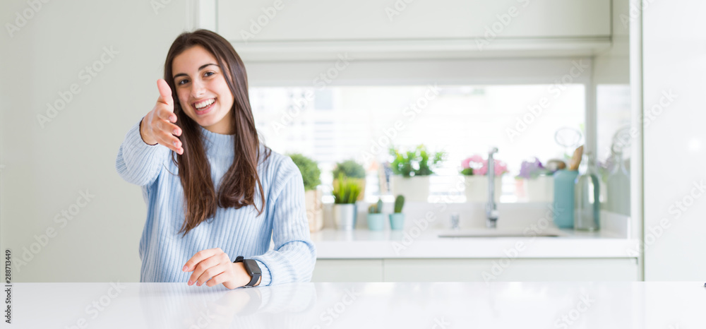 Wide angle picture of beautiful young woman sitting on white table at home smiling friendly offering handshake as greeting and welcoming. Successful business. Wide angle picture of beautiful young woman sitting on white table at home smiling friendly offering handshake as greeting and welcoming. Successful business.