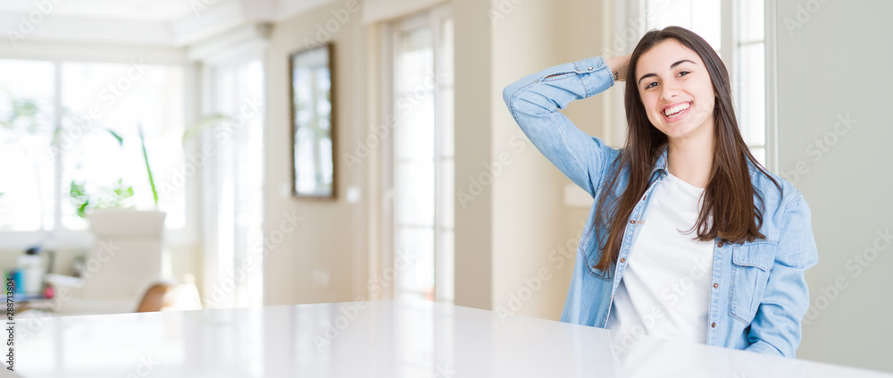 Wide angle picture of beautiful young woman sitting on white table at home Smiling confident touching hair with hand up gesture, posing attractive Wide angle picture of beautiful young woman sitting on white table at home Smiling confident touching hair with hand up gesture, posing attractive