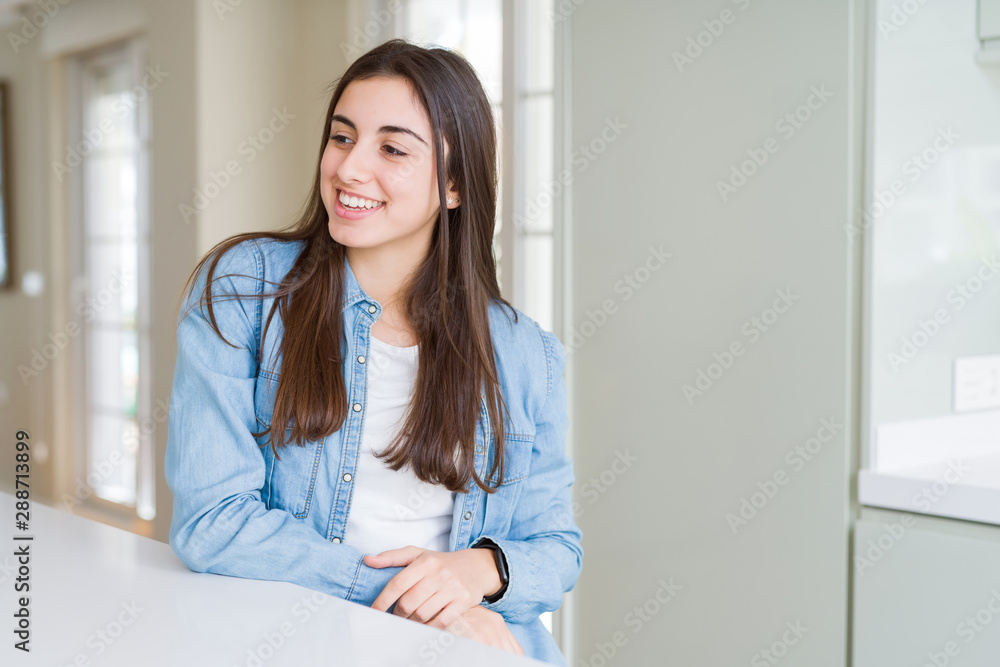 Beautiful young woman sitting on white table at home looking away to side with smile on face, natural expression. Laughing confident. Beautiful young woman sitting on white table at home looking away to side with smile on face, natural expression. Laughing confident.