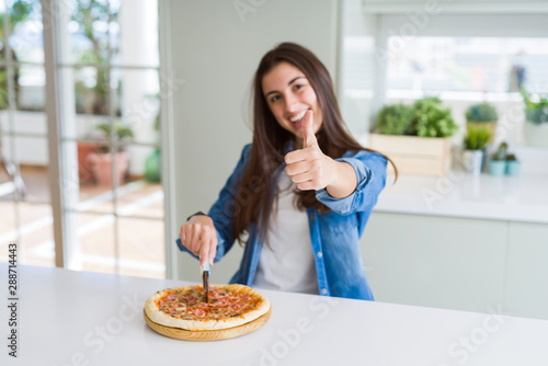 Beautiful young woman cutting a tasty pizza slice using a cutter happy with big smile doing ok sign, thumb up with fingers, excellent sign