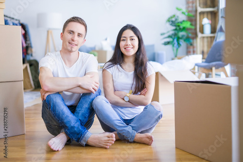 Young beautiful couple sitting on the floor at new home around cardboard boxes happy face smiling with crossed arms looking at the camera. Positive person.