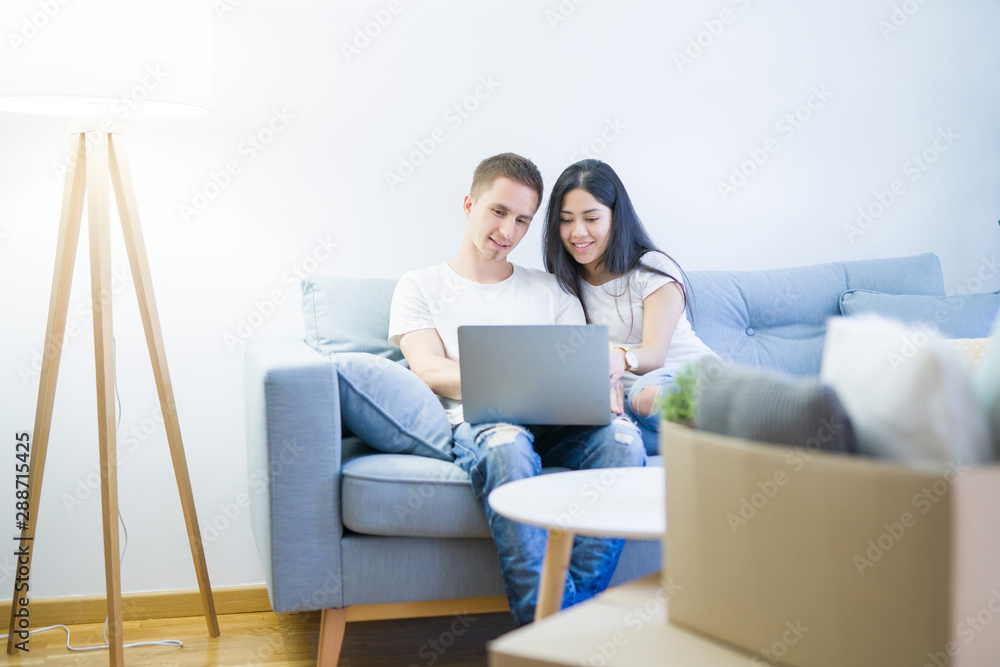 Young beautiful couple sitting on the sofa using laptop at new home around cardboard boxes