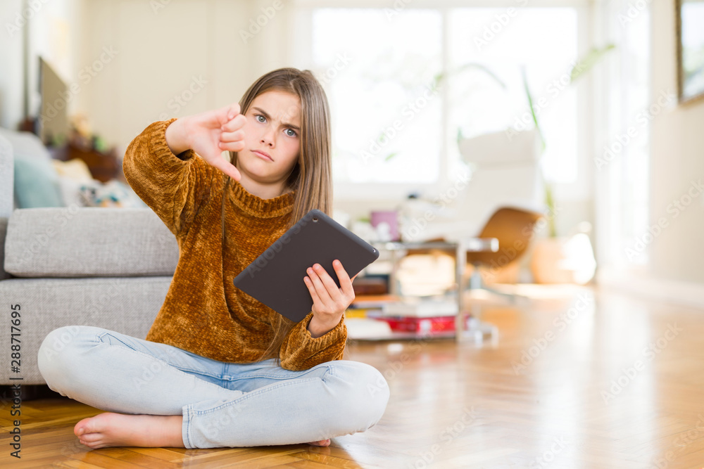 Beautiful young girl kid using digital touchpad tablet sitting on the floor with angry face, negative sign showing dislike with thumbs down, rejection concept Beautiful young girl kid using digital touchpad tablet sitting on the floor with angry face, negative sign showing dislike with thumbs down, rejection concept