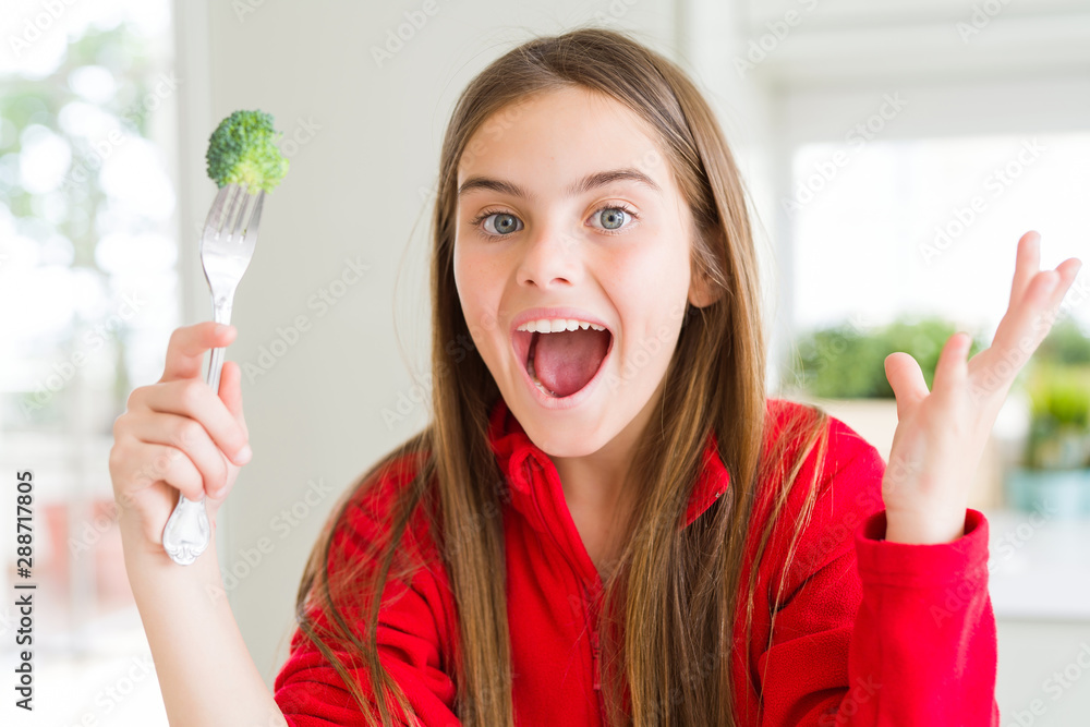 Beautiful young girl eating fresh broccoli very happy and excited, winner expression celebrating victory screaming with big smile and raised hands Beautiful young girl eating fresh broccoli very happy and excited, winner expression celebrating victory screaming with big smile and raised hands