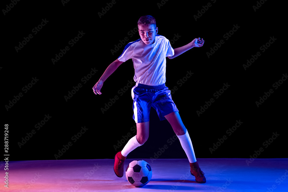 Young boy as a soccer or football player in sportwear making a feint or a kick with the ball for a goal on dark studio background. Fit playing boy in action, movement, motion at game. Purple neon
