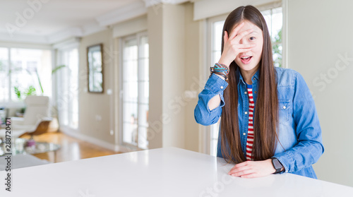 Young beautiful asian woman with long hair wearing denim jacket peeking in shock covering face and eyes with hand  looking through fingers with embarrassed expression.