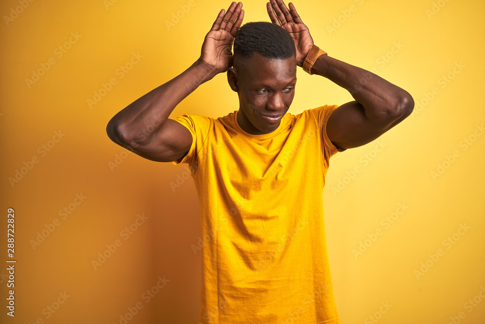 Young african american man wearing casual t-shirt standing over isolated yellow background Doing bunny ears gesture with hands palms looking cynical and skeptical. Easter rabbit concept. Young african american man wearing casual t-shirt standing over isolated yellow background Doing bunny ears gesture with hands palms looking cynical and skeptical. Easter rabbit concept.