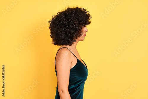 Young arab woman with curly hair wearing elegant dress over isolated yellow background looking to side, relax profile pose with natural face with confident smile.