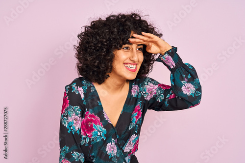 Quadro su tela Young arab woman with curly hair wearing floral dress over isolated pink background very happy and smiling looking far away with hand over head