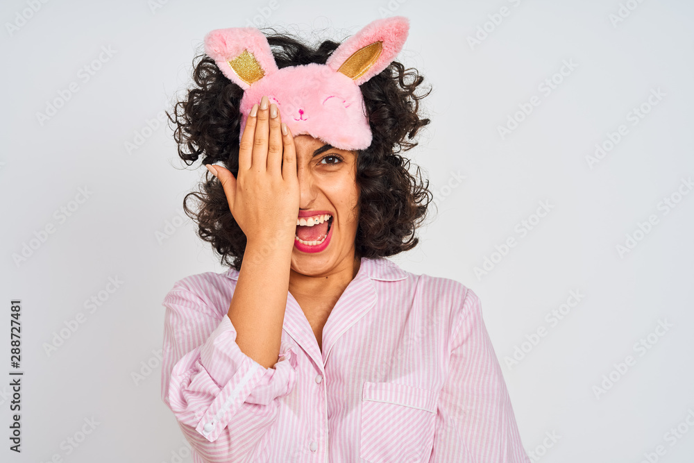 Arab woman with curly hair wearing pajama and sleep mask over isolated white background covering one eye with hand, confident smile on face and surprise emotion.