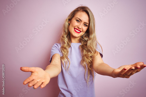 Young beautiful woman wearing purple t-shirt standing over pink isolated background looking at the camera smiling with open arms for hug. Cheerful expression embracing happiness.